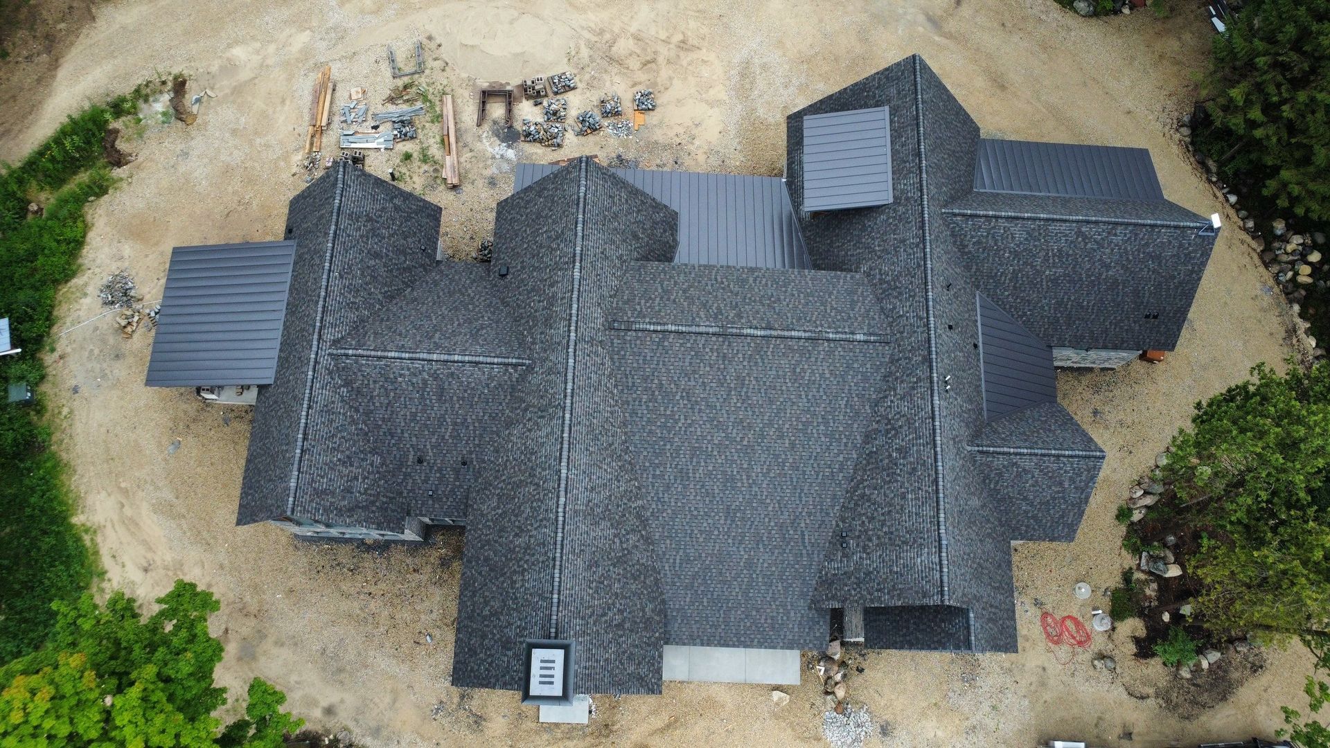 Aerial top-down view of a complex, dark-shingled residential roof surrounded by unfinished dirt and surrounding trees.