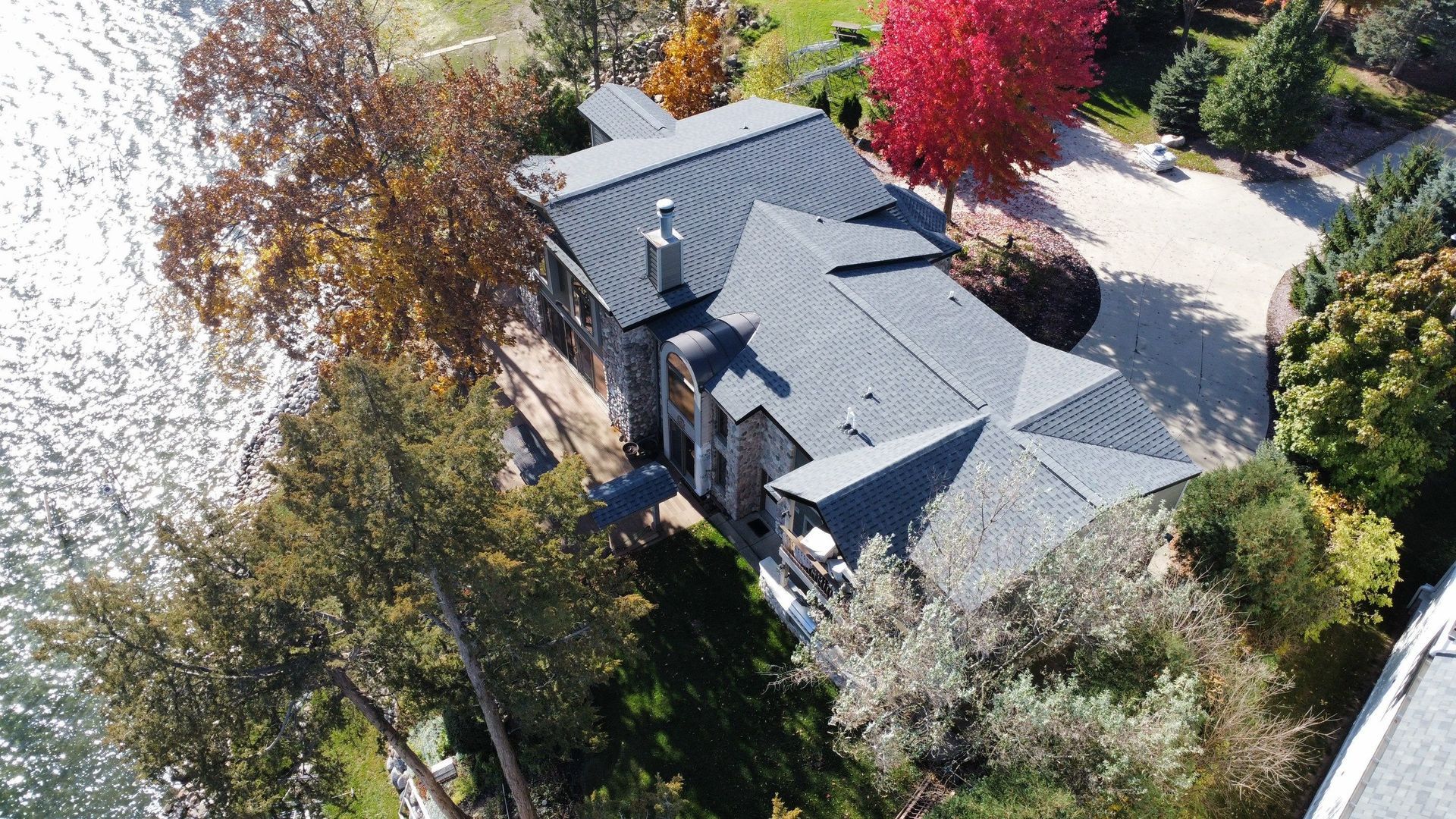 Aerial view of a gray-roofed stone house nestled among colorful autumn trees next to a sparkling lake.