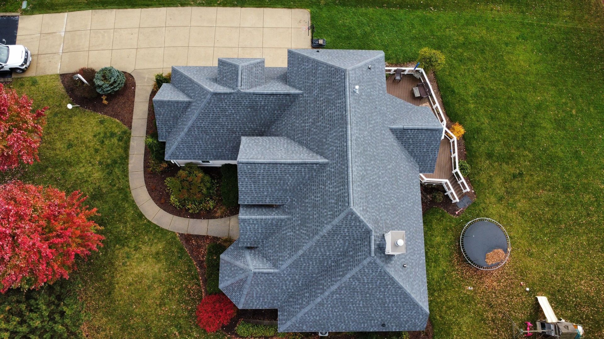An aerial view of a gray shingled residential roof, surrounded by a lawn, a concrete driveway, and some autumn trees.