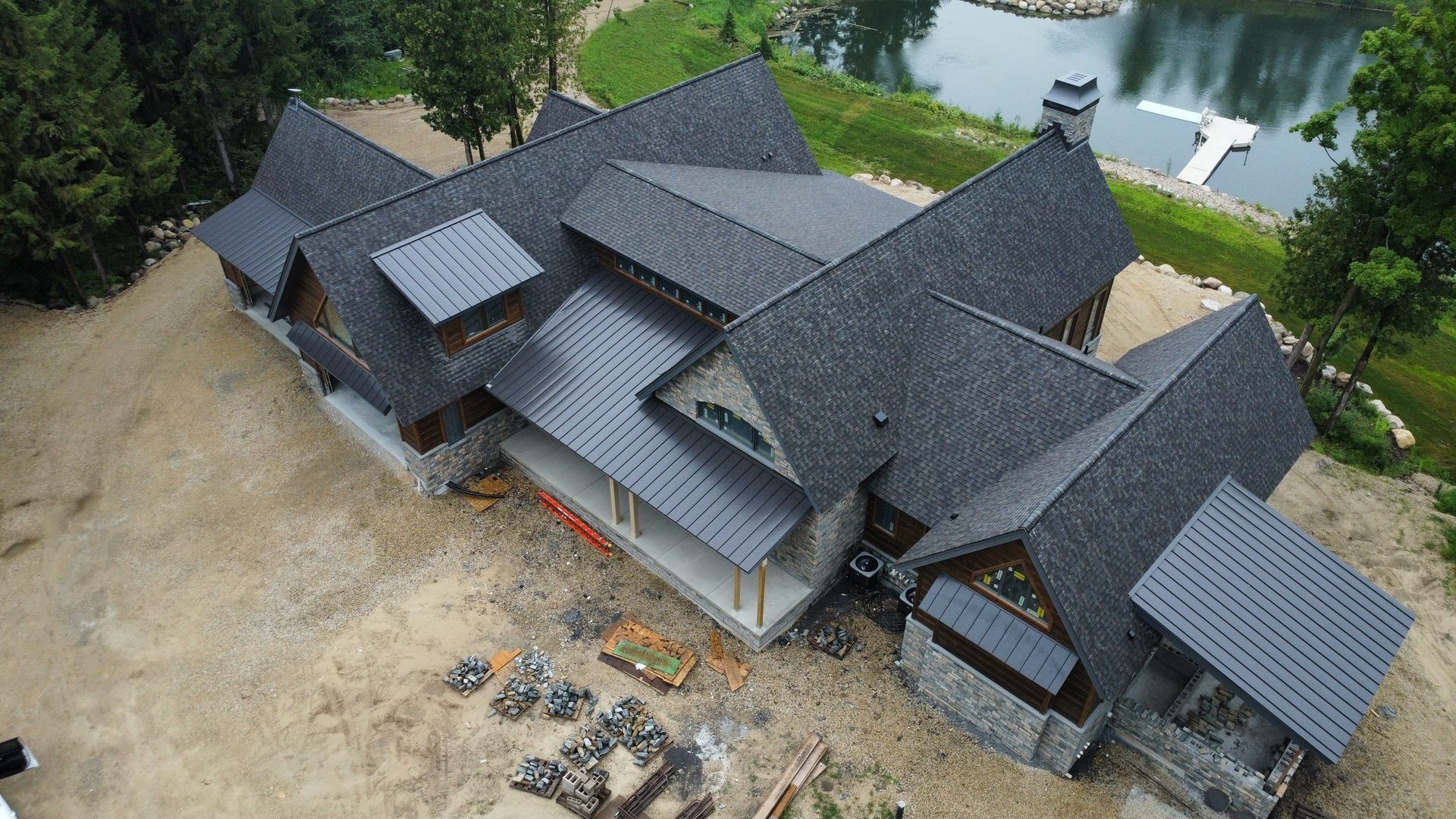 An aerial view of a stone and timber home with a dark shingled and metal roof, located on a lot near a small pond.