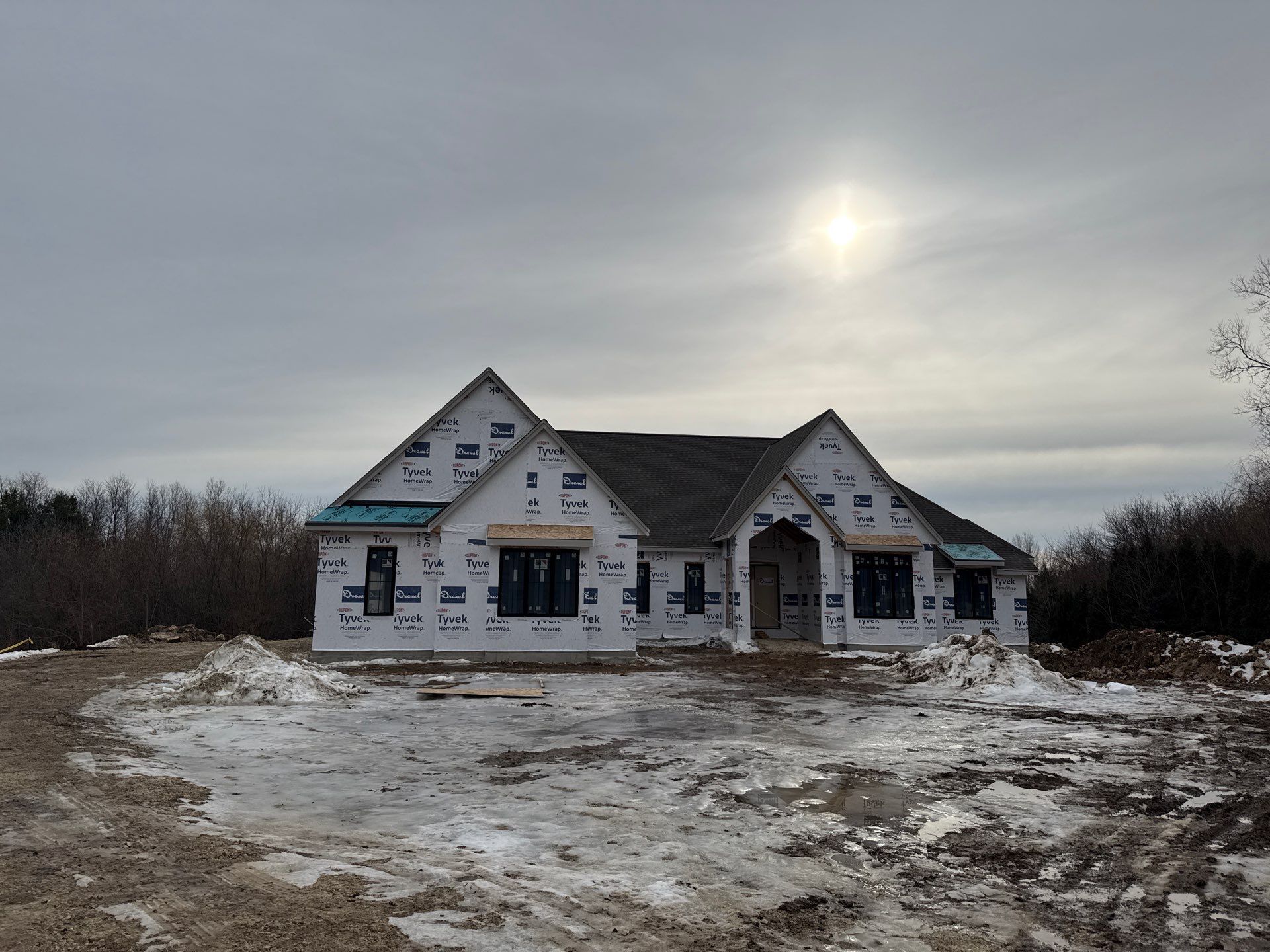 A new house under construction with white exterior sheathing, black window frames, and a gray roof, surrounded by snow.