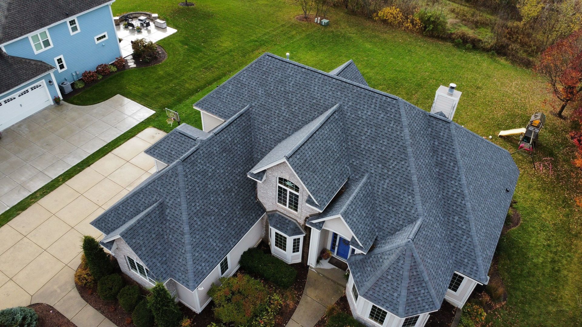Aerial view of a suburban house with a gray shingled roof, a paved driveway, and a surrounding lawn with trees.