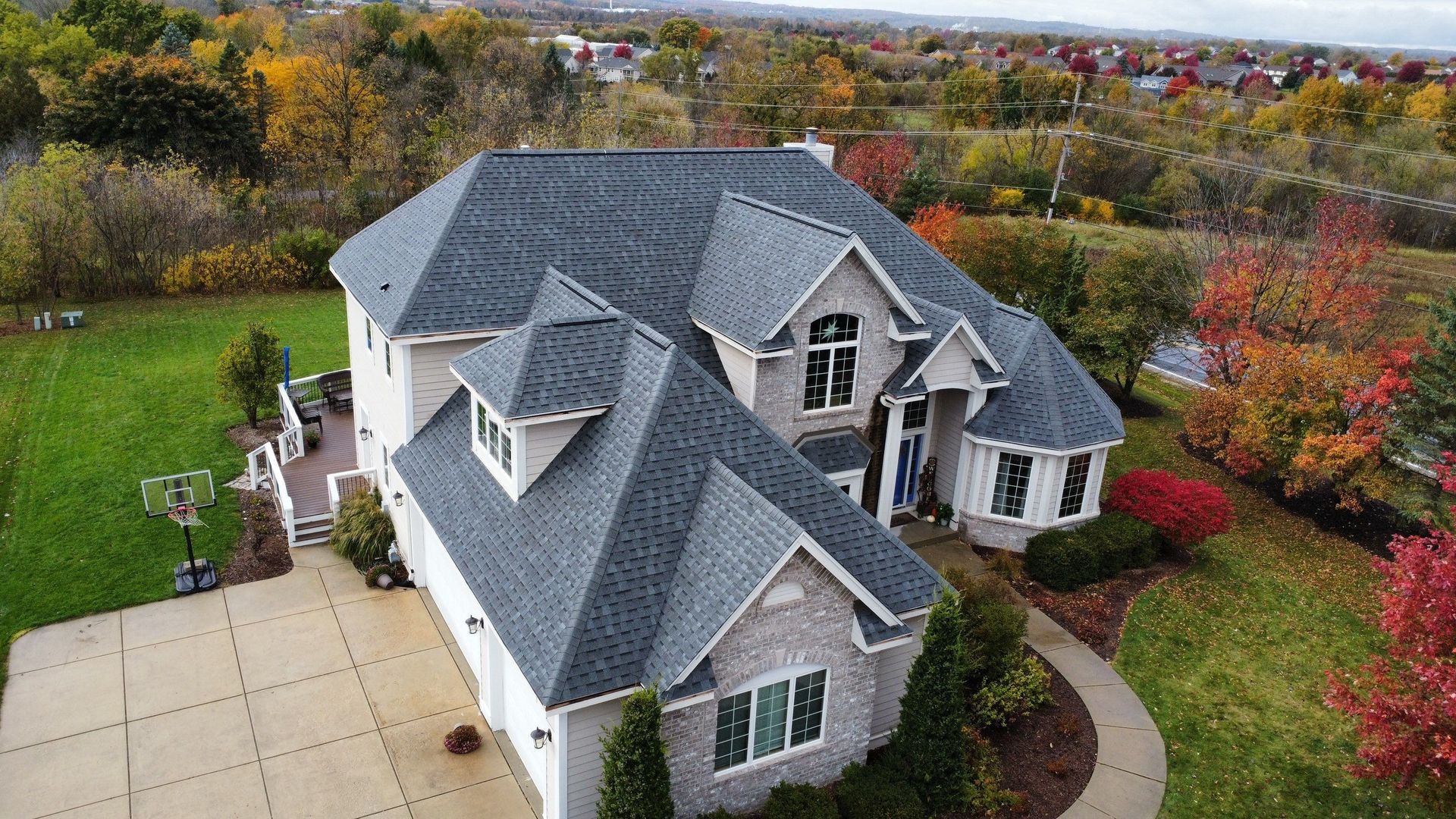 Aerial view of a suburban brick house with a gray shingled roof, surrounded by trees with autumn foliage.