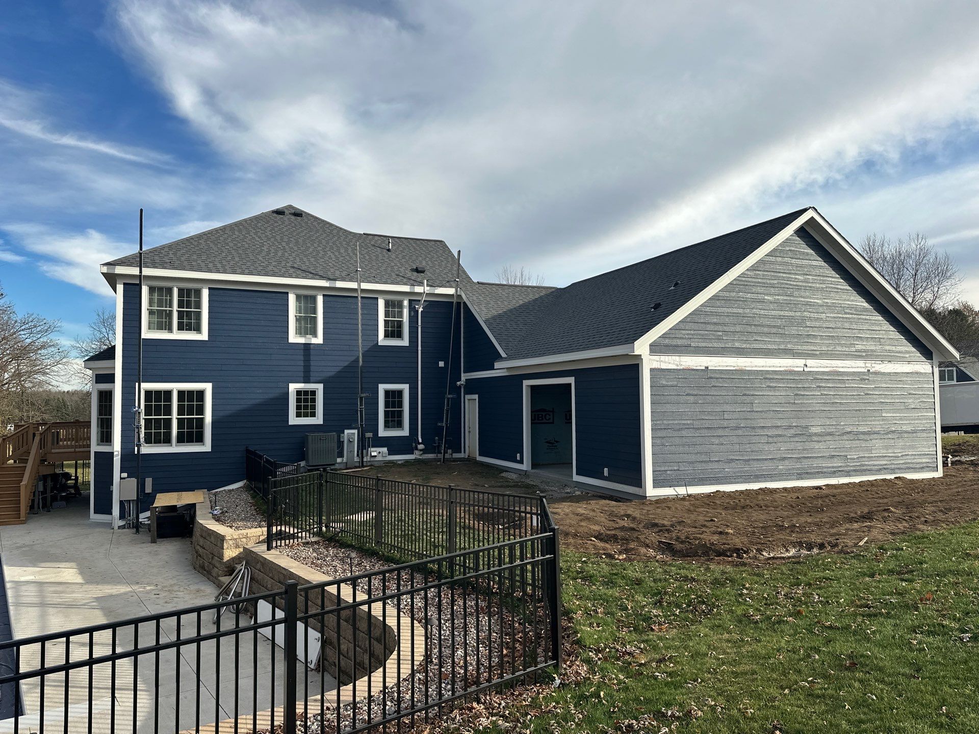 A two-story blue house with a matching garage addition, featuring a stone retaining wall and black metal fencing outdoors.