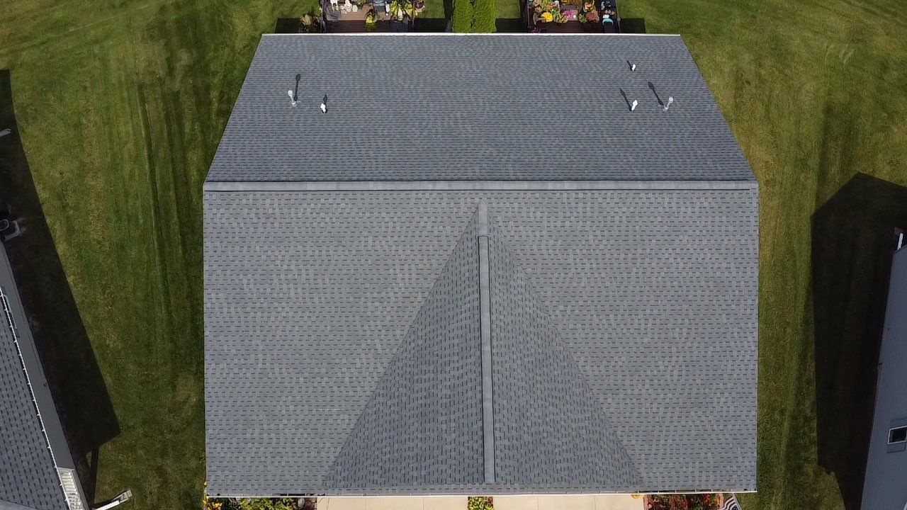 An overhead, eye-level view of a dark gray shingled roof with a central ridge, showing a square section against grass.