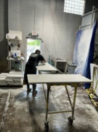 Person sanding a long, light-colored surface on a rolling table inside a workshop.