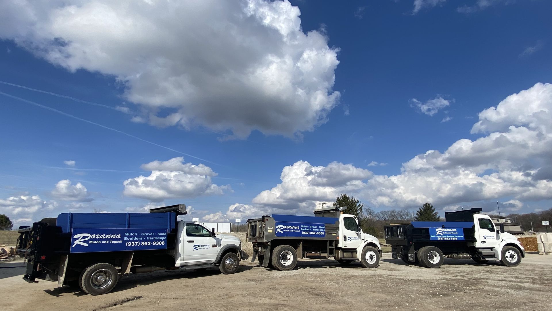 Three white dump trucks with blue beds parked in a gravel lot under a bright cloudy sky.