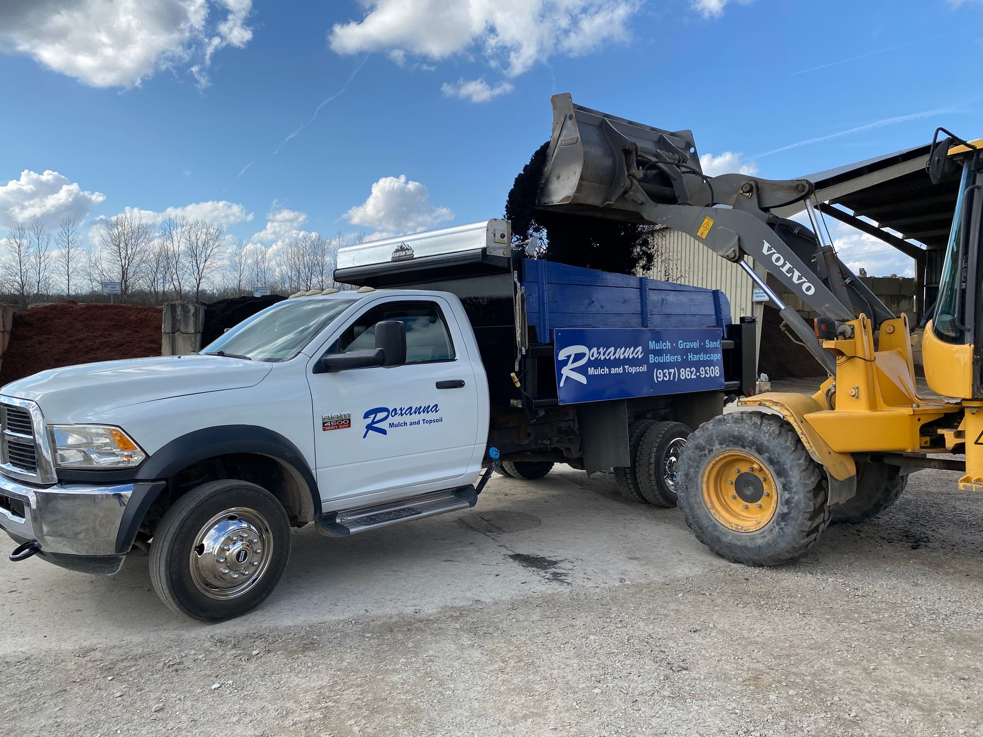 White pickup truck beside yellow front loader dumping dirt in a gravel yard under a blue sky