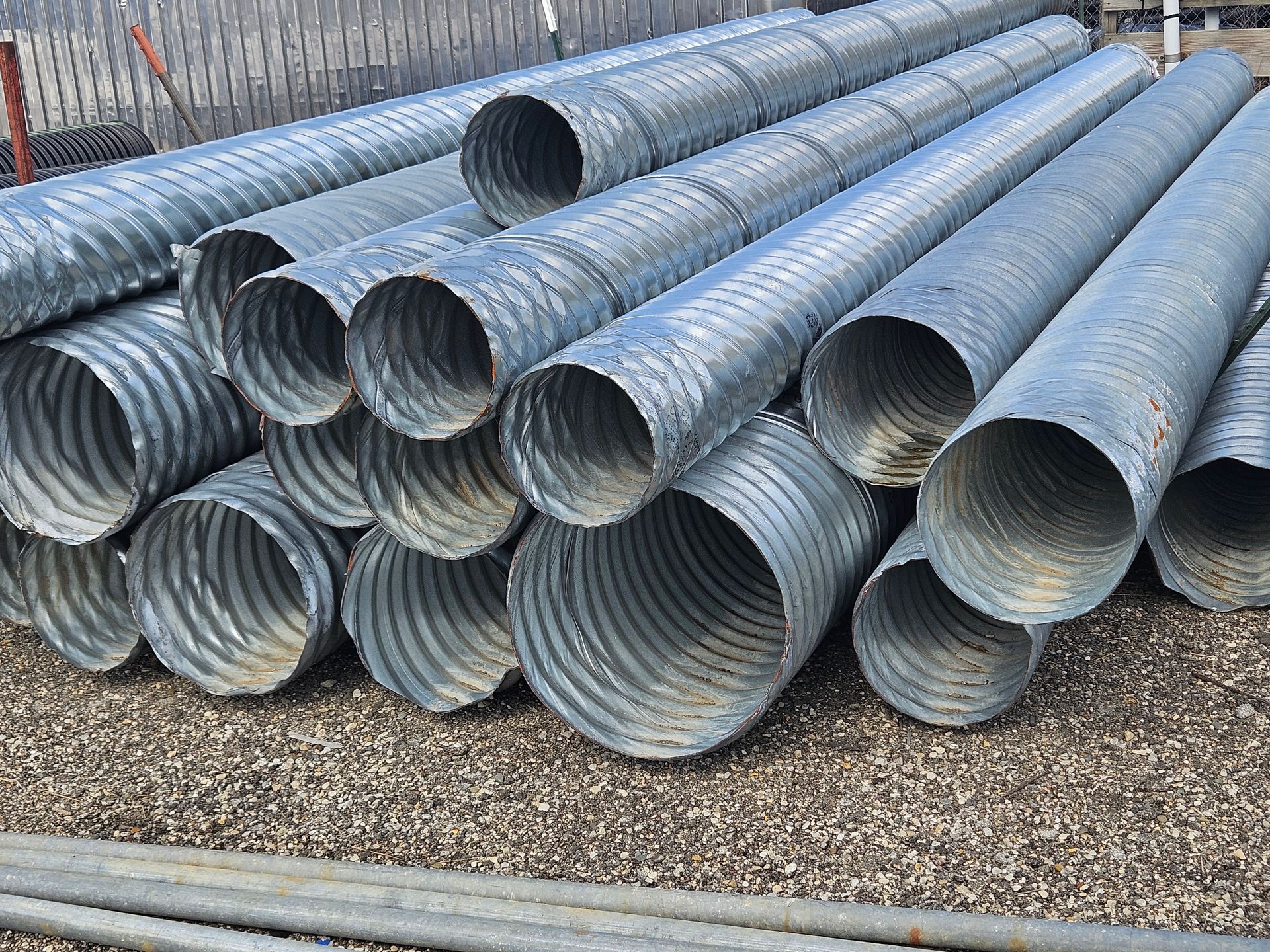 Stacked gray corrugated metal pipes on gravel in an outdoor storage yard