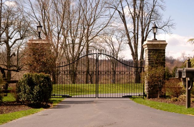 Black iron gate between brick pillars at the entrance to a tree-lined driveway or estate.