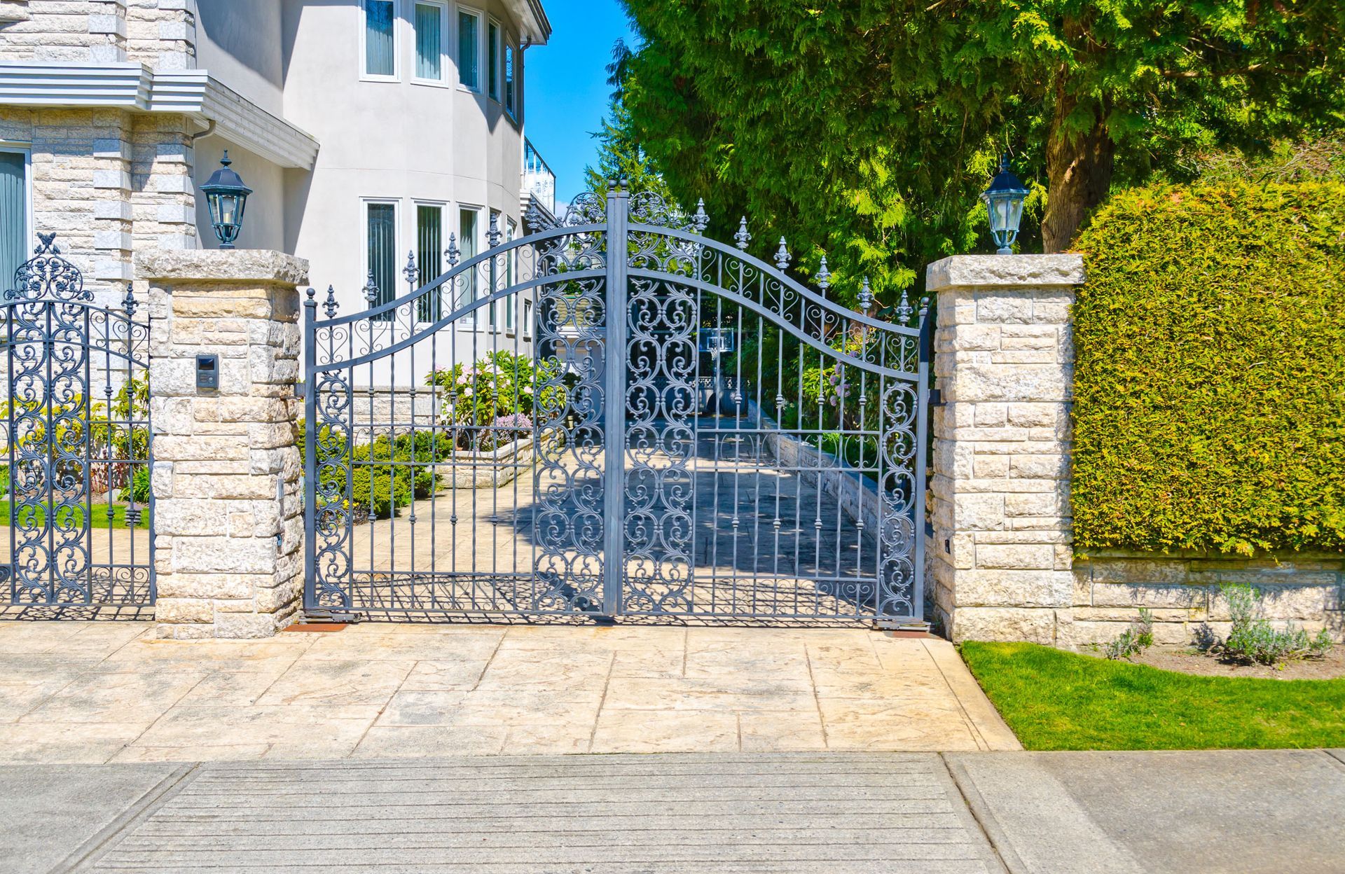 Ornate metal driveway gate between stone pillars and hedges in front of a white house