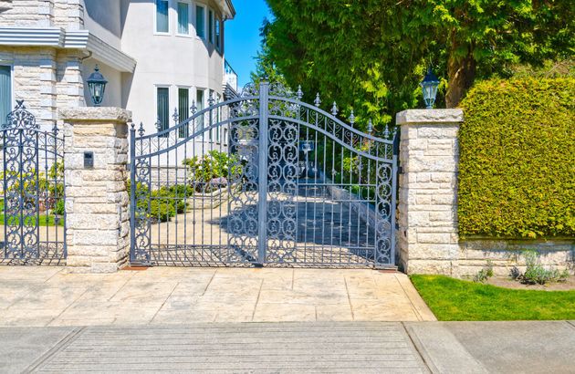 Ornate metal driveway gate between stone pillars and hedges in front of a white house