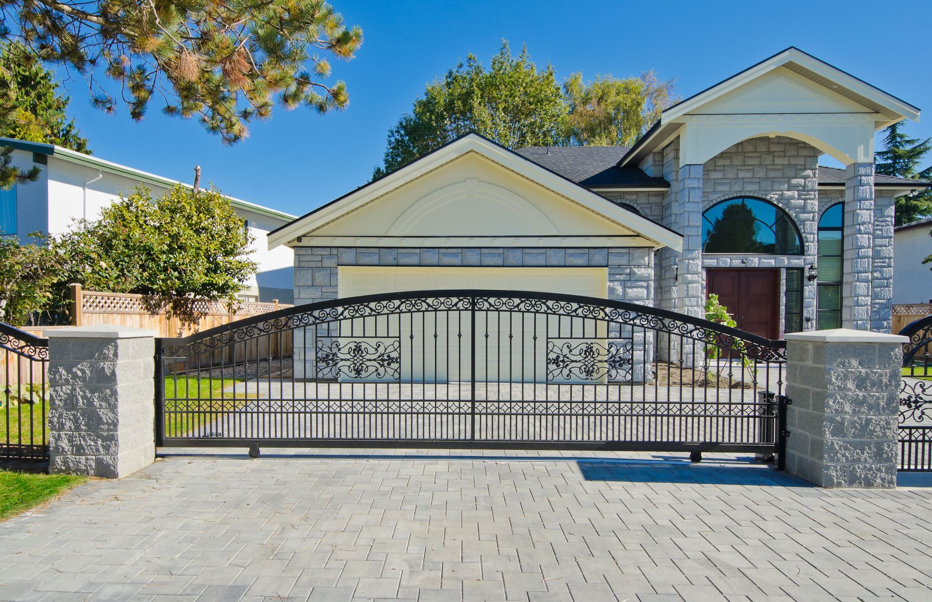 Large gated suburban house with stone columns, arched entry, and paved driveway under a blue sky