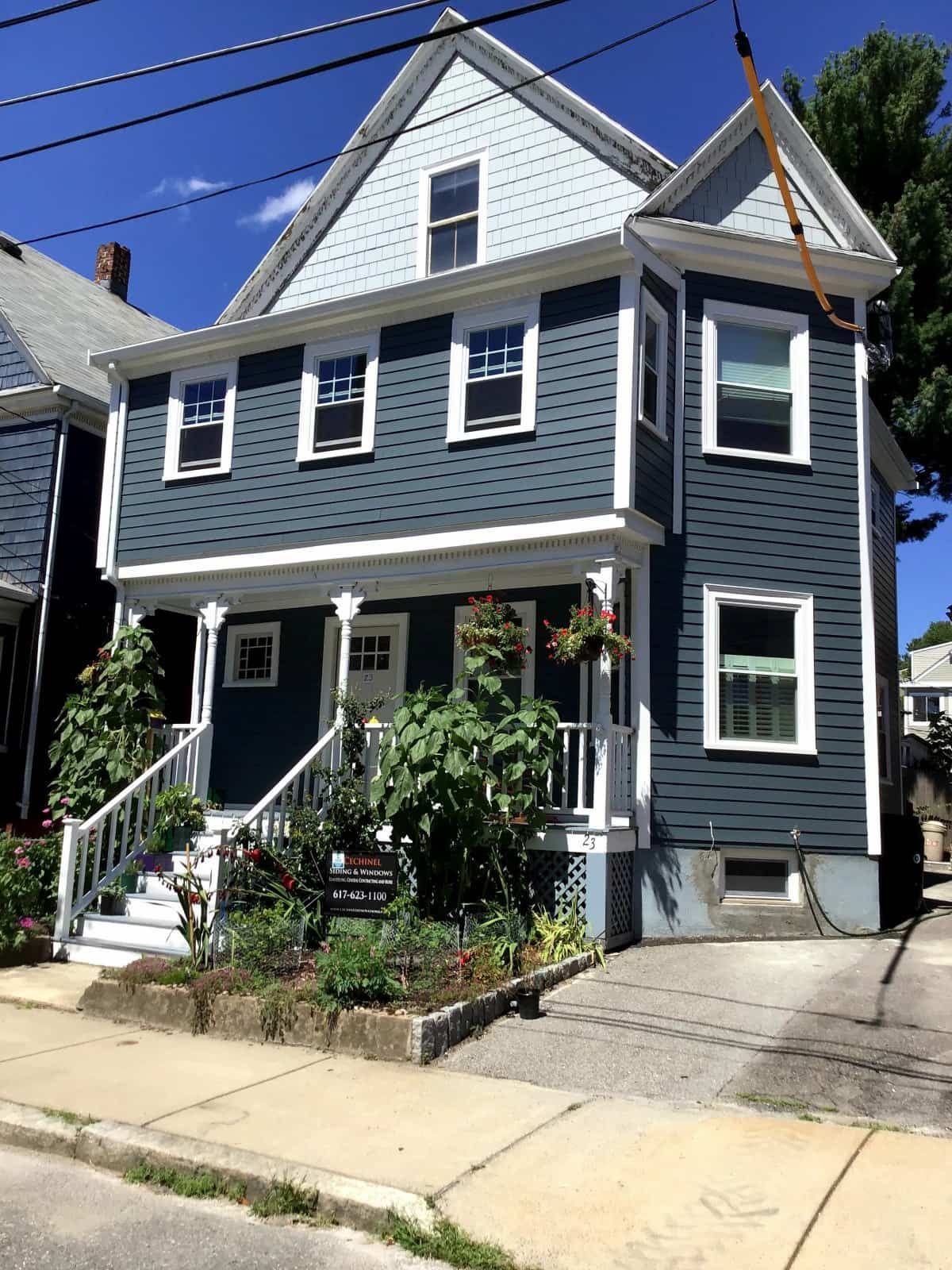 Two-story light green house with white porch, triangular roof, and a small tower. Orange porta-potty in front.