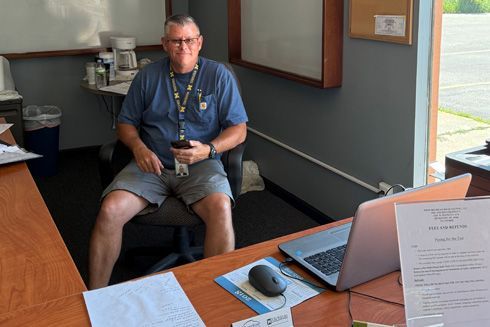 Man sitting at desk, wearing glasses, blue shirt, gray shorts, holding phone, in office.