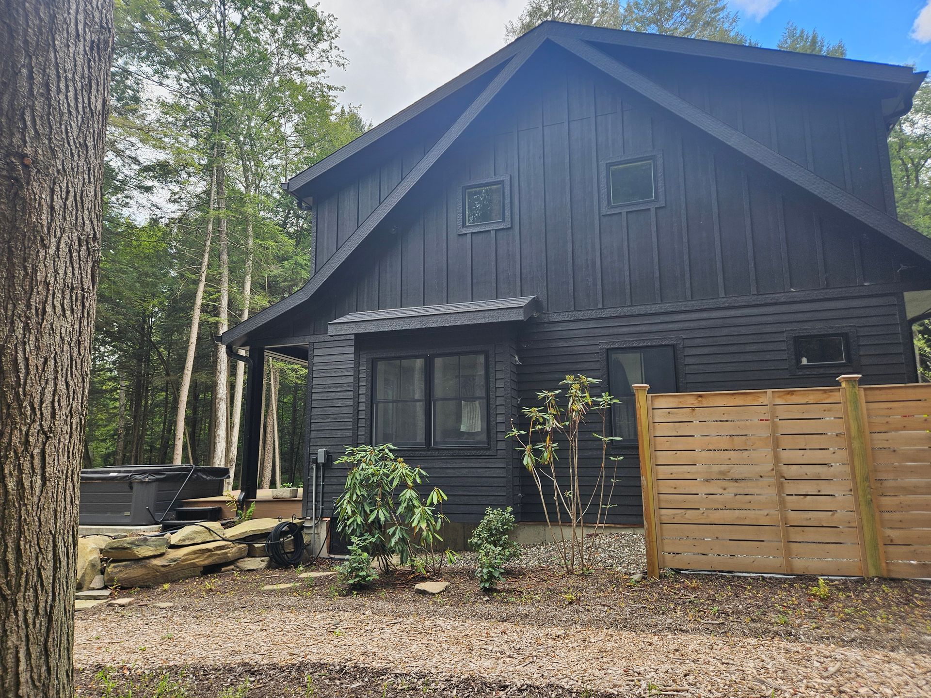 A modern, two-story, dark-sided cabin nestled in a wooded area with a wooden privacy fence and a nearby hot tub.