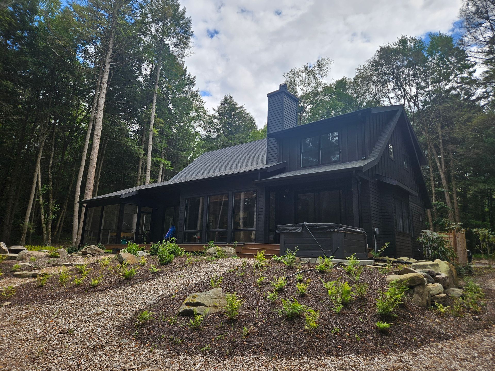 A black cabin with a metal roof and a stone chimney, set in a wooded area with a gravel path and landscaped garden.