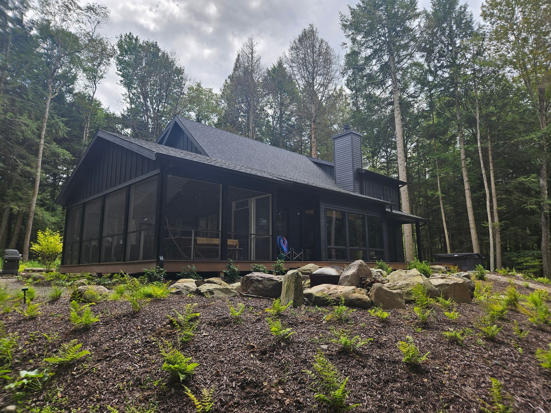 A black modern cabin with a screened-in porch, surrounded by lush green trees and a rock-landscaped yard.