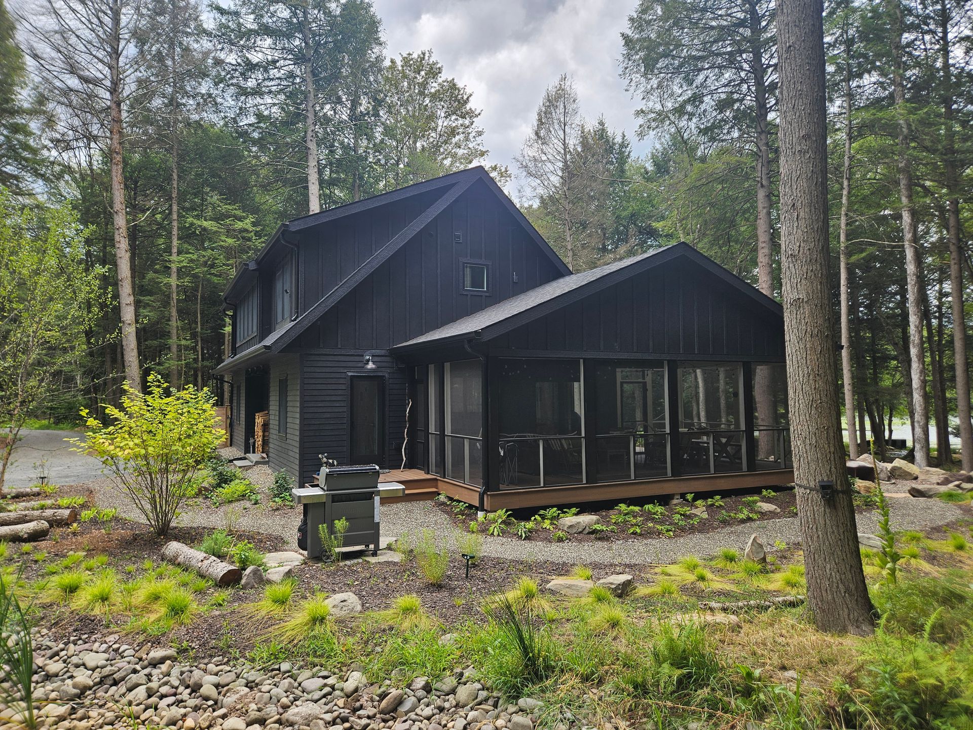 A modern black, two-story house with a screened-in porch, surrounded by lush trees and a stone-covered yard.