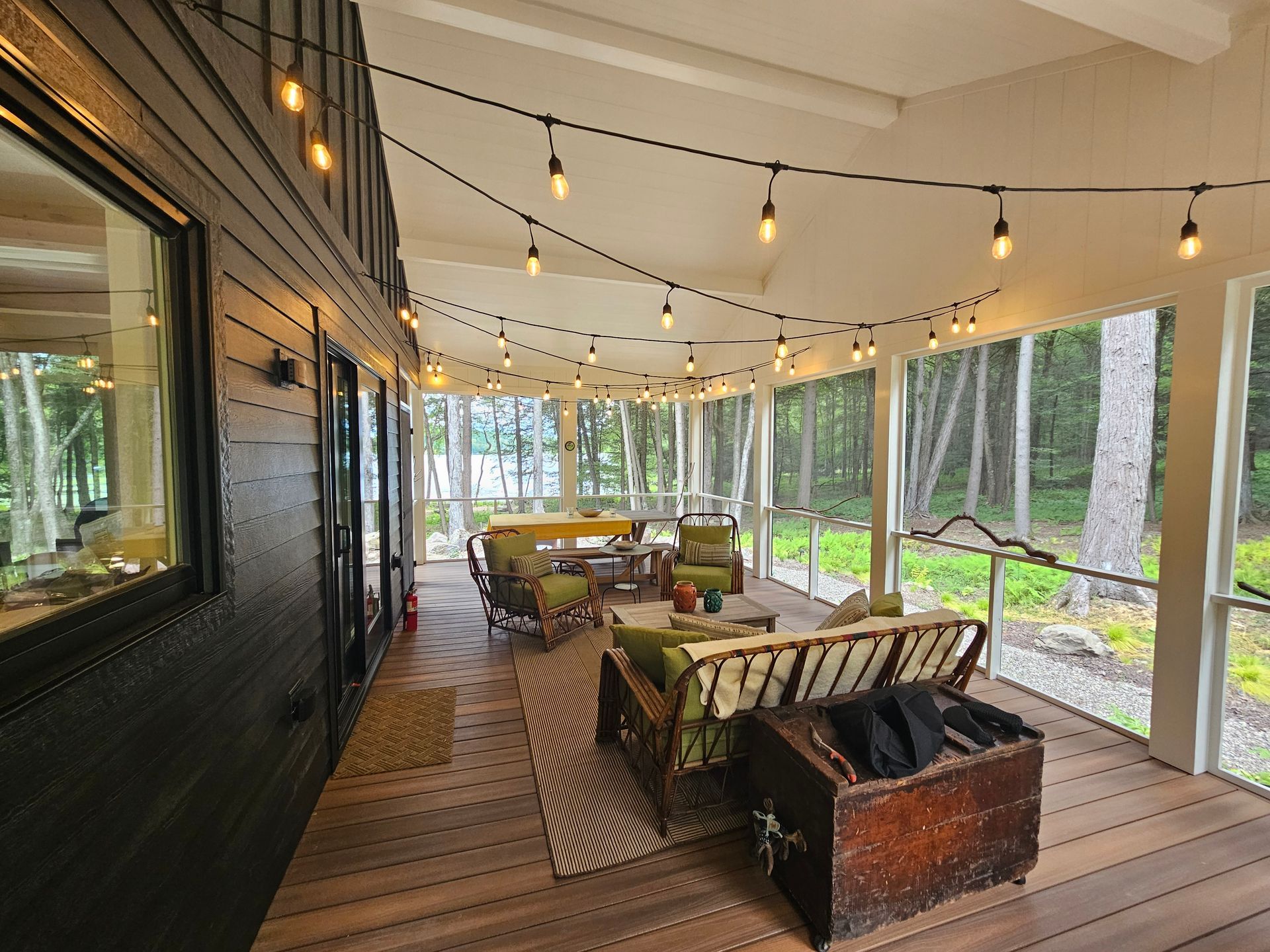 A covered wooden porch features patio furniture and string lights, overlooking a treed landscape.