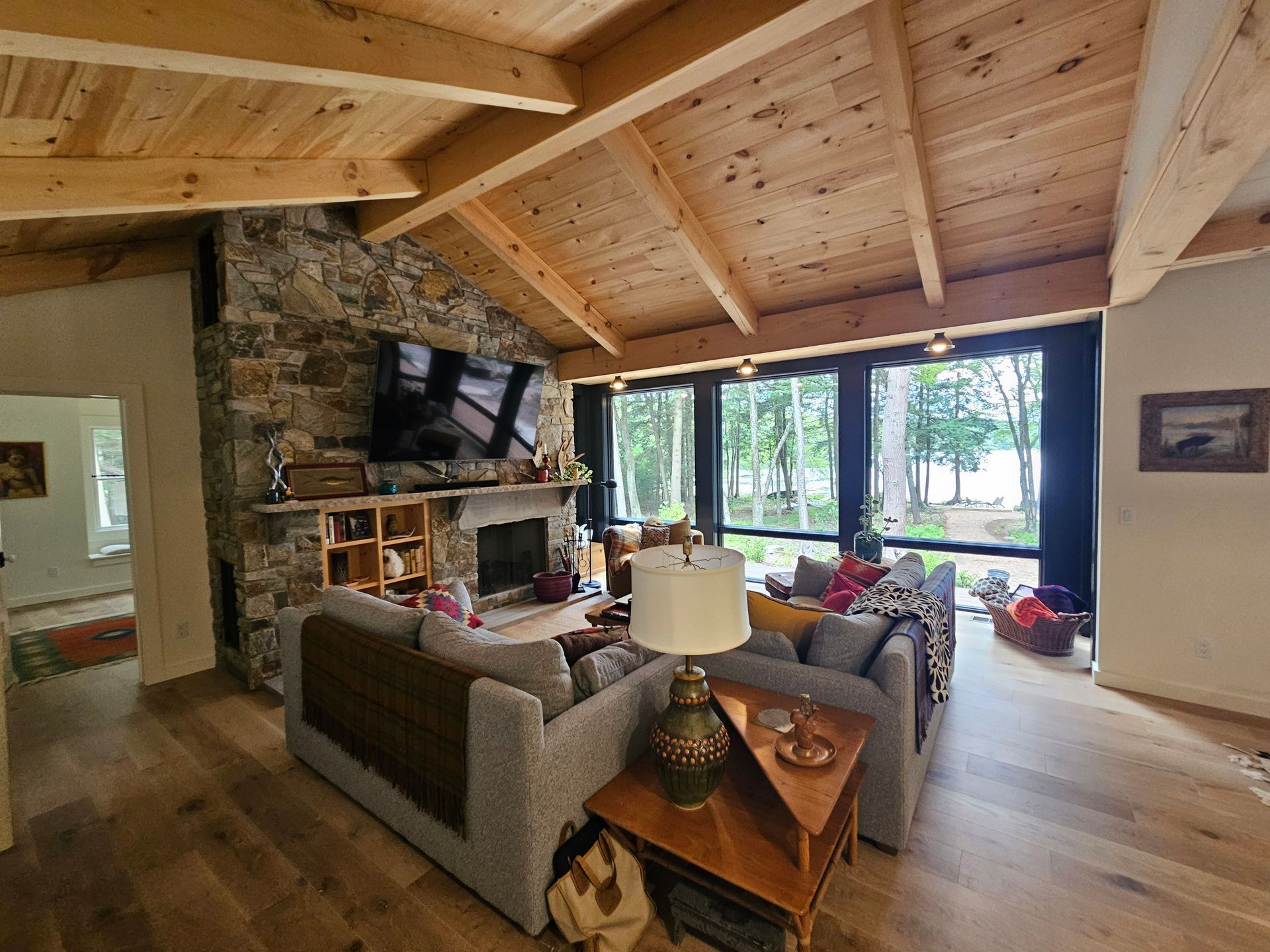 Living room with a stone fireplace, exposed wooden beams, and a large gray sectional sofa facing a windowed wall.