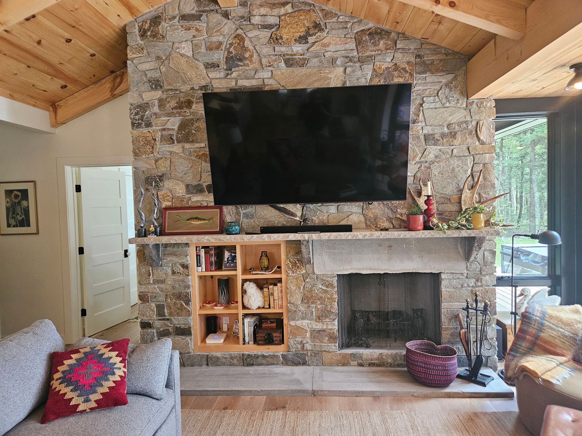 A stone fireplace with a mounted television, built-in shelving, and a grey sofa in a rustic living room with wooden beams.