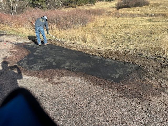 Man raking asphalt on a road patch, outdoors, surrounded by grass and brush.