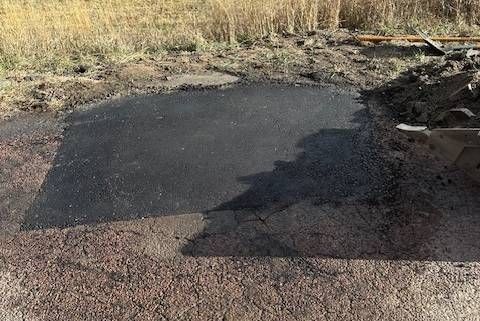 Patch of fresh asphalt on a dirt and gravel surface, near dry grass and some debris.