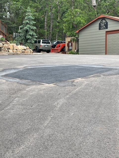 Asphalt driveway with freshly paved patch; vehicles and garage in background.