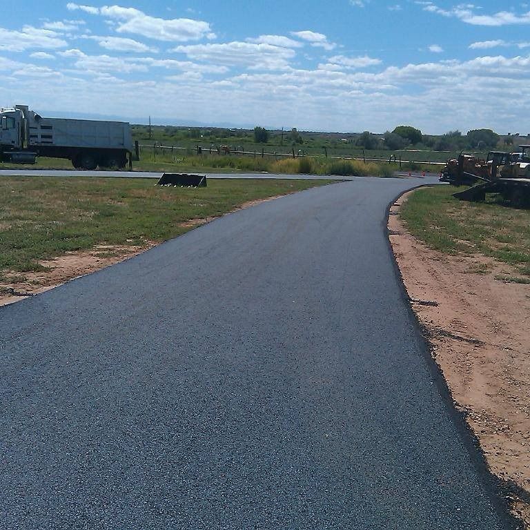 Asphalt road curving through a grassy area, with construction equipment and truck visible in the background under a blue sky.