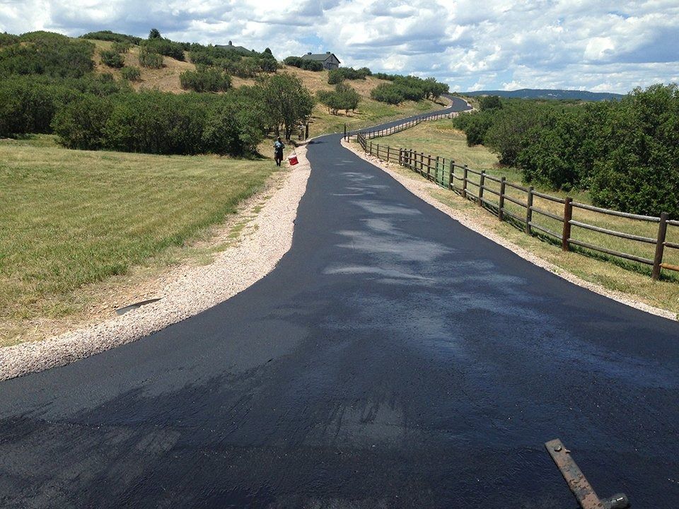 Newly paved asphalt driveway curves uphill through a grassy landscape, next to a wooden fence.