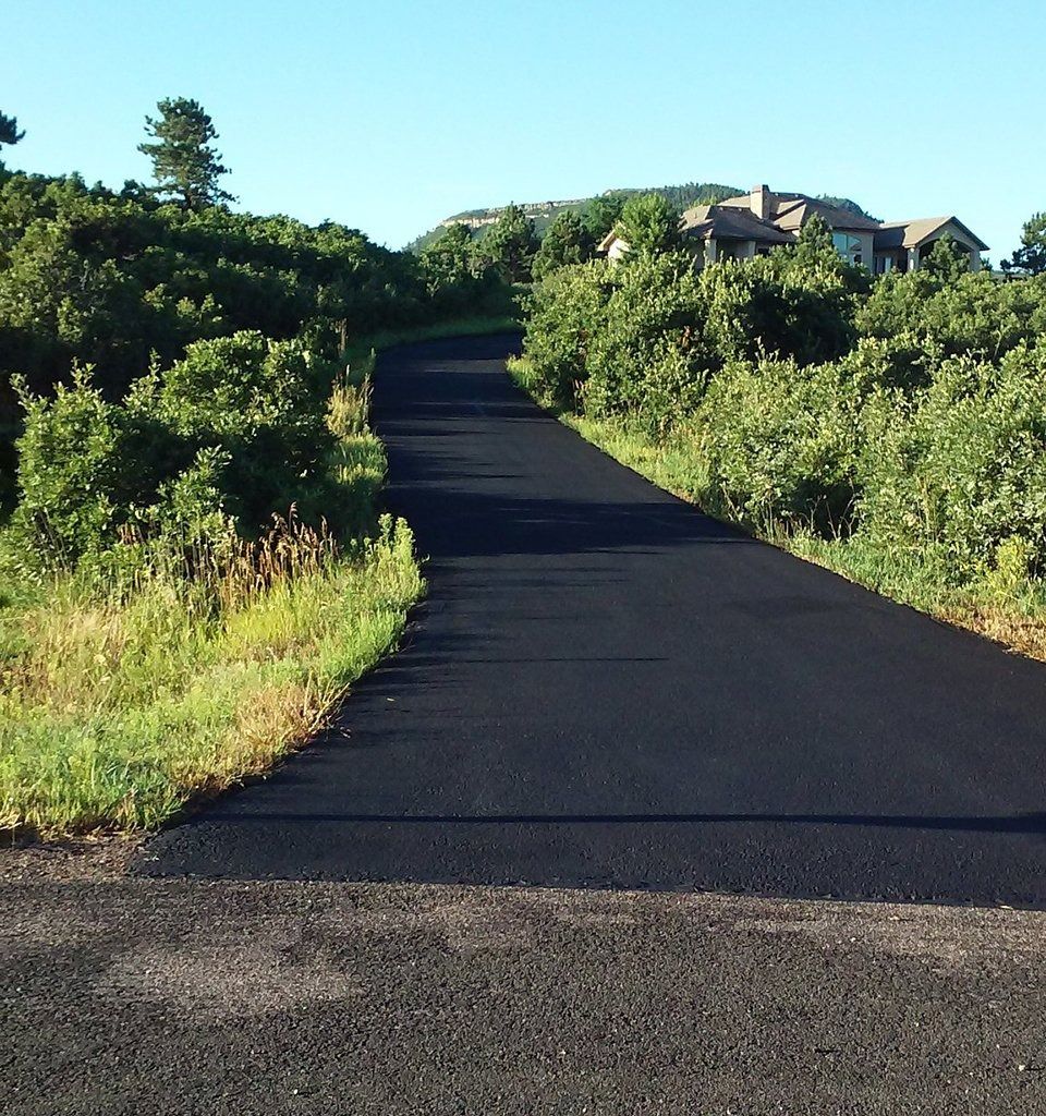 Black asphalt driveway winding uphill through greenery toward a house under a blue sky.