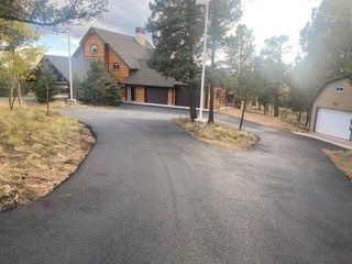 Paved driveway leading to a large wood-sided house and detached garage in a wooded setting.