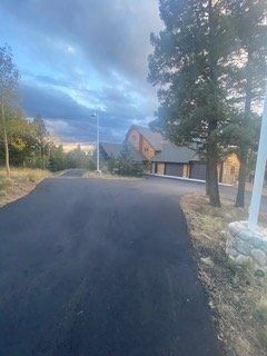 Paved driveway leading to a large wood house under a cloudy sky. Trees and a stone base fence marker are visible.