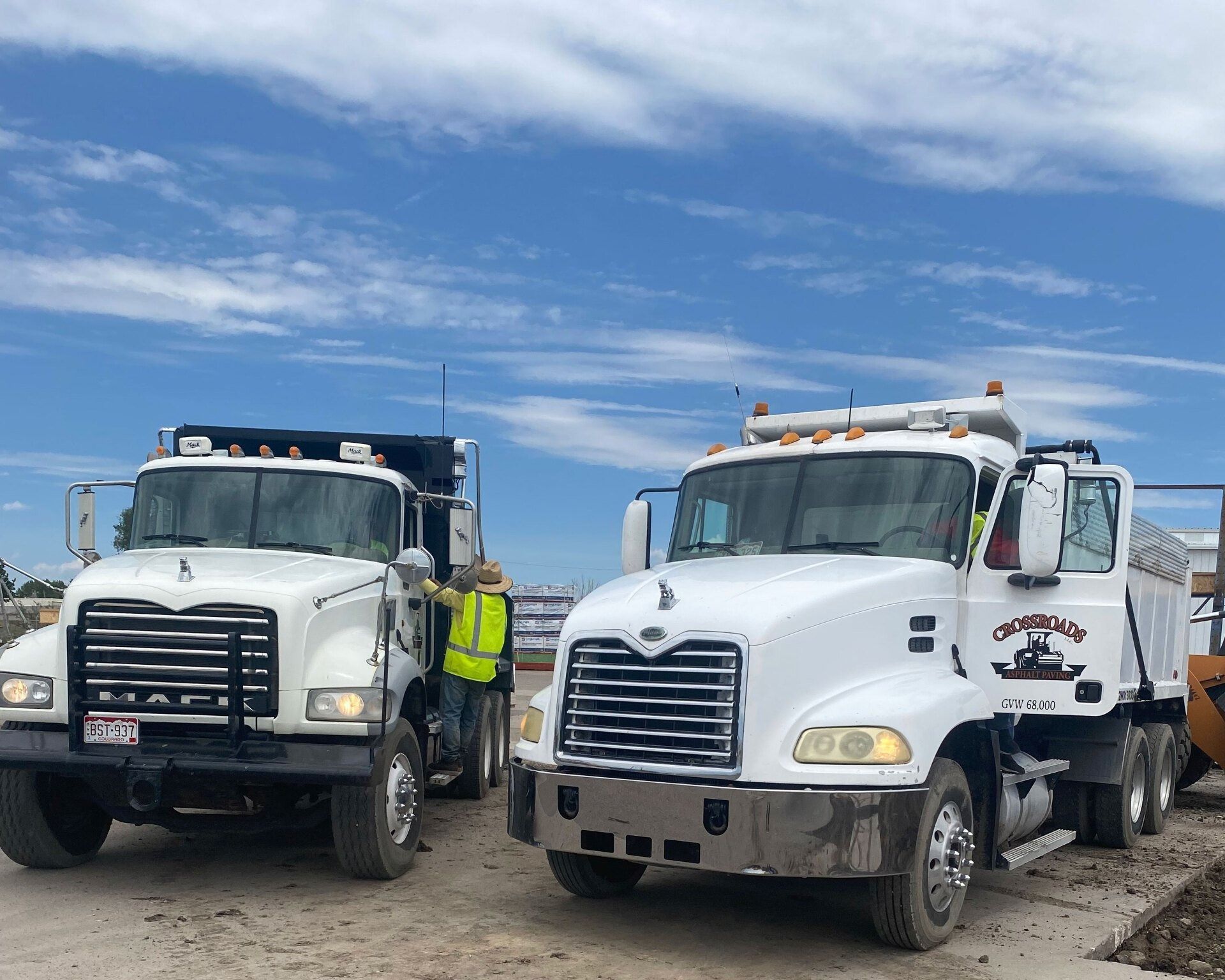 Two white dump trucks parked at a construction site. Workers in vests stand near trucks under a blue sky.