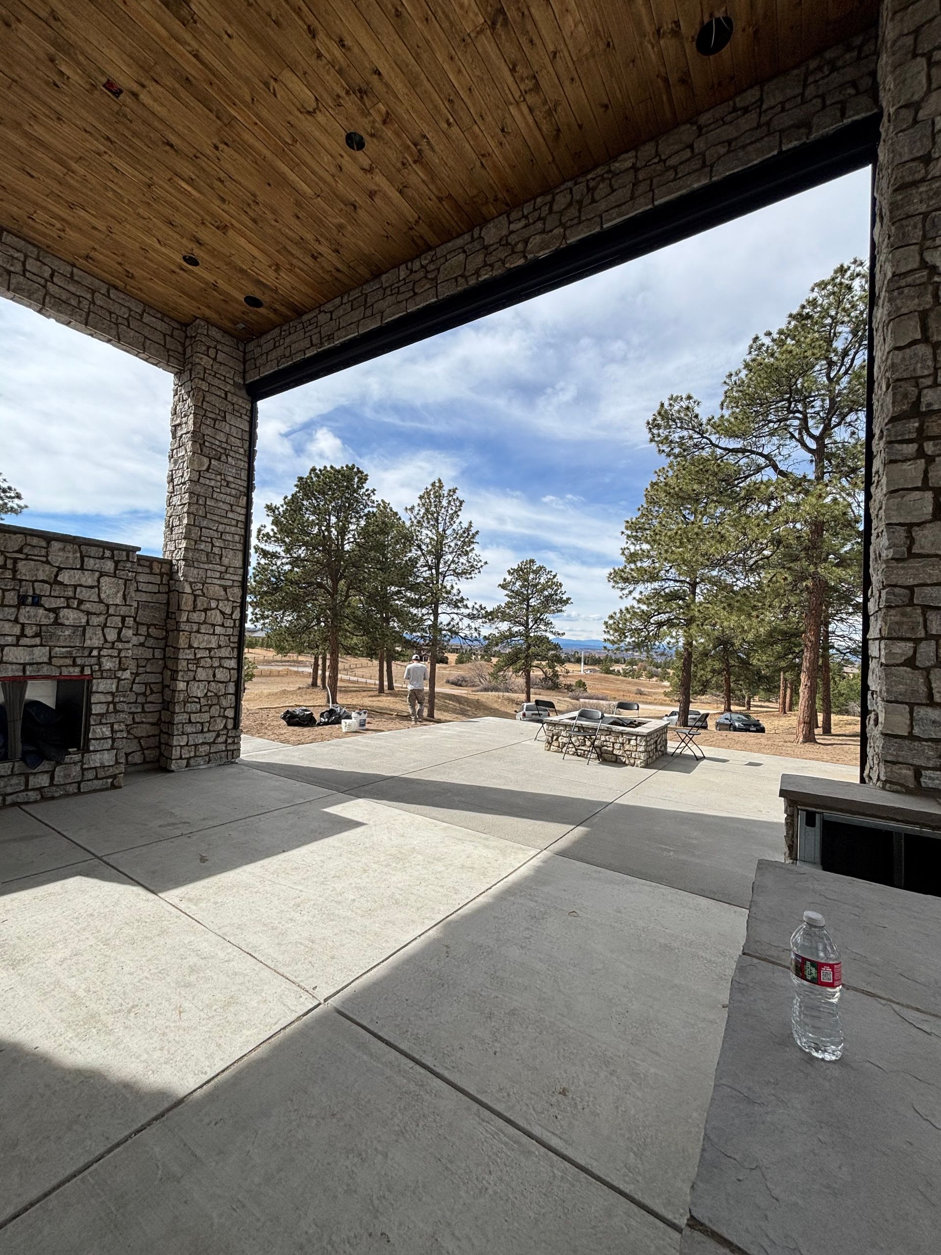 Covered patio with stone fireplace overlooking a pine-filled landscape under a cloudy sky.