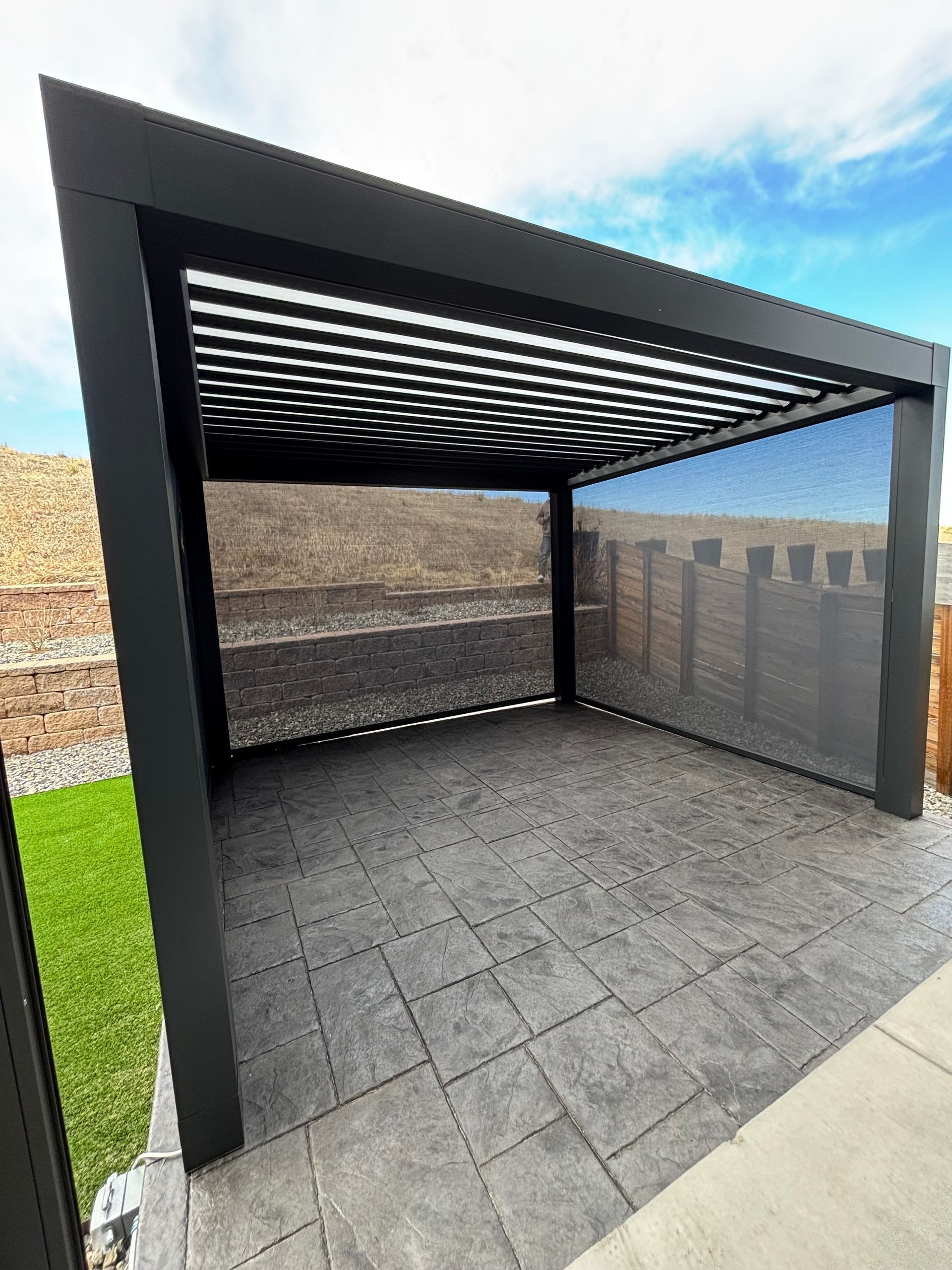 Black pergola with adjustable roof and side screen on a stone patio.