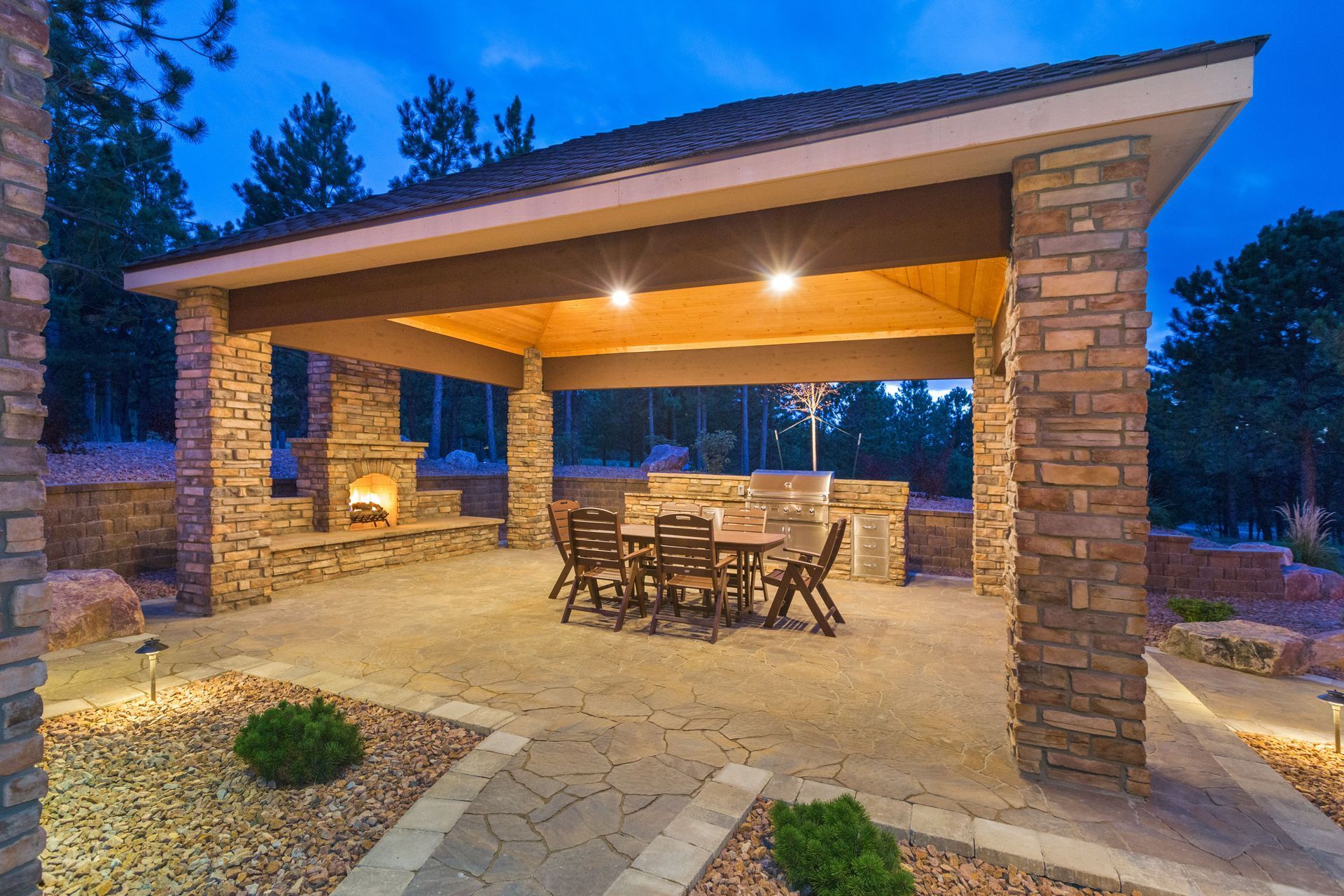 Covered outdoor patio with stone pillars, lit ceiling, and dining table at dusk