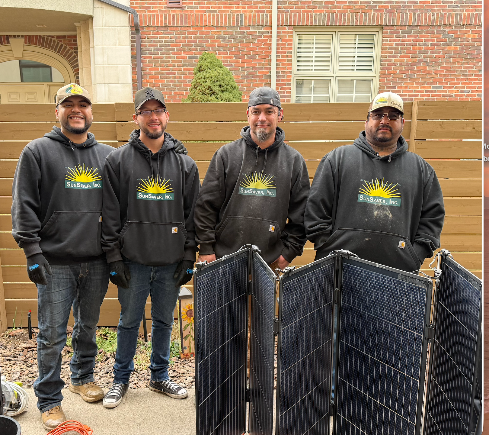 Four people in black hoodies standing behind folded black panels in a backyard.
