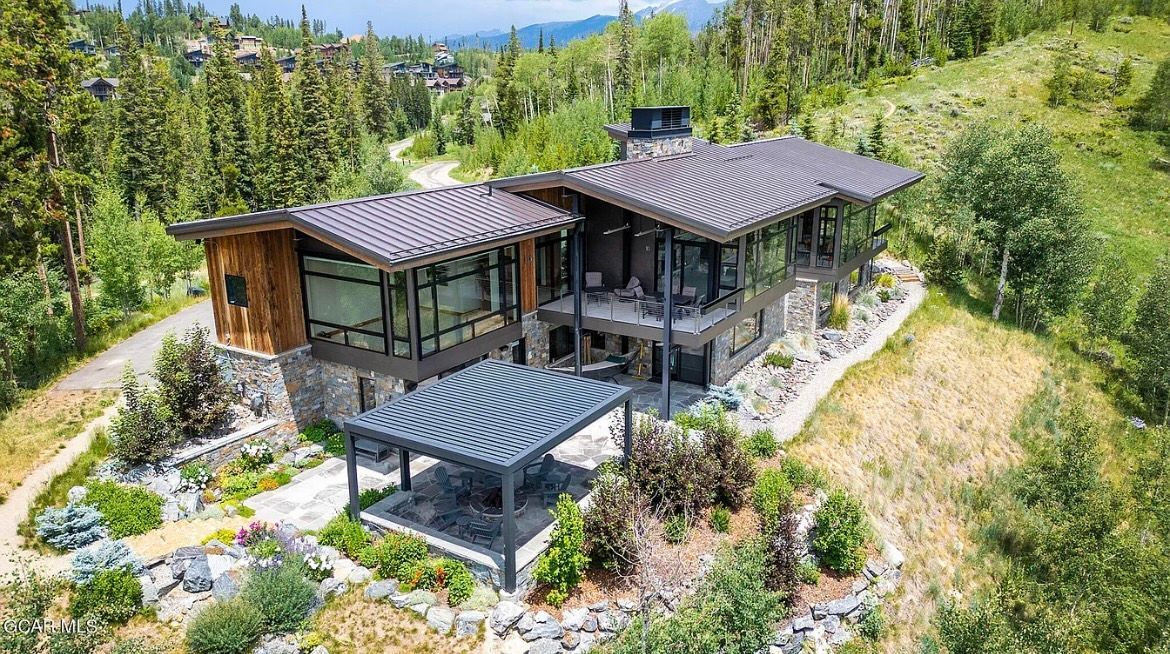 Modern hillside house with dark roof, large windows, and landscaped garden surrounded by trees