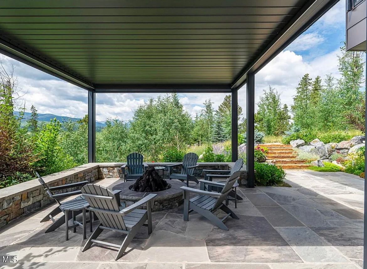 Covered patio with wooden chairs, fire pit table, stone flooring, and a garden view