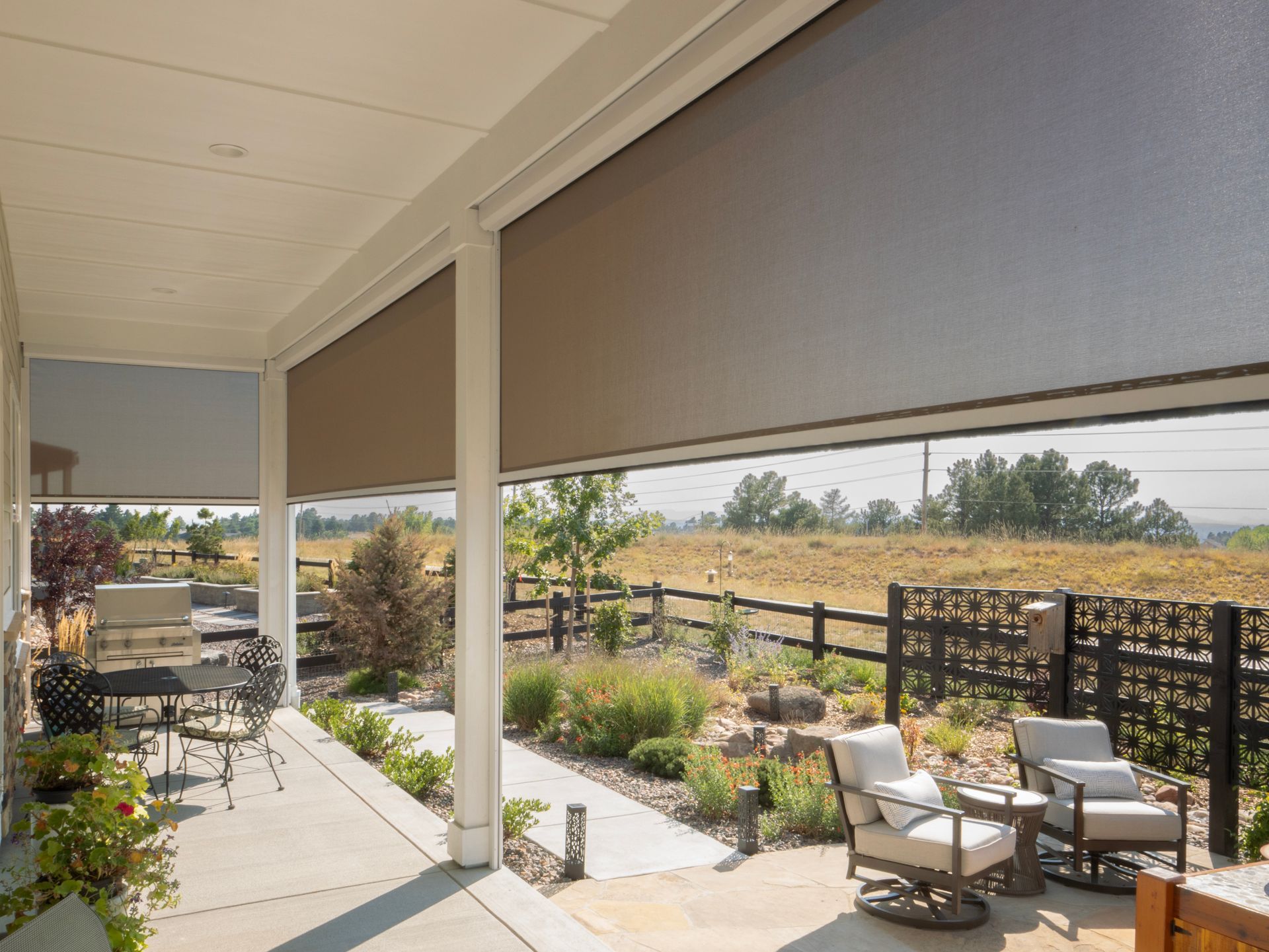 Covered patio with outdoor seating, planters, and rolled-down shades overlooking a landscaped yard