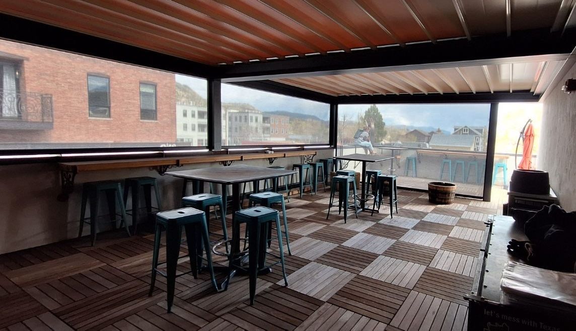 Open-air rooftop café with empty stools, checkered floor, and city view under a wooden canopy