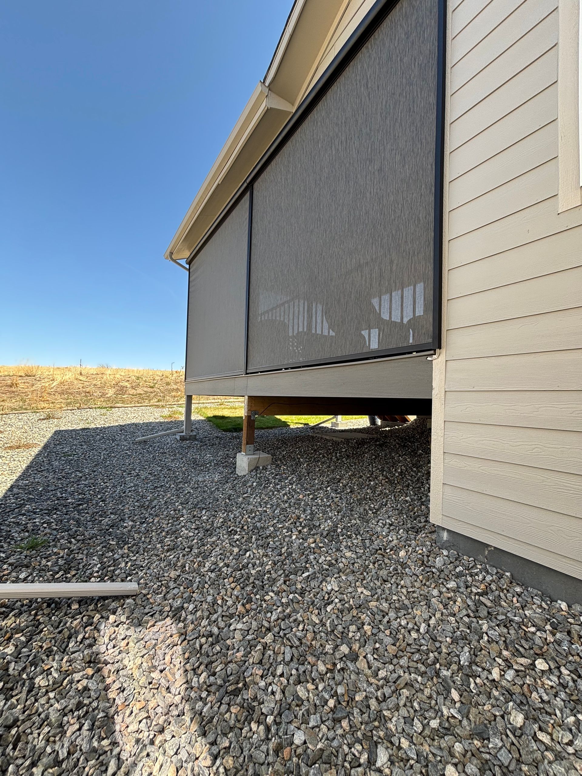 Raised house with black screened porch over gravel beside a beige wall under a clear blue sky