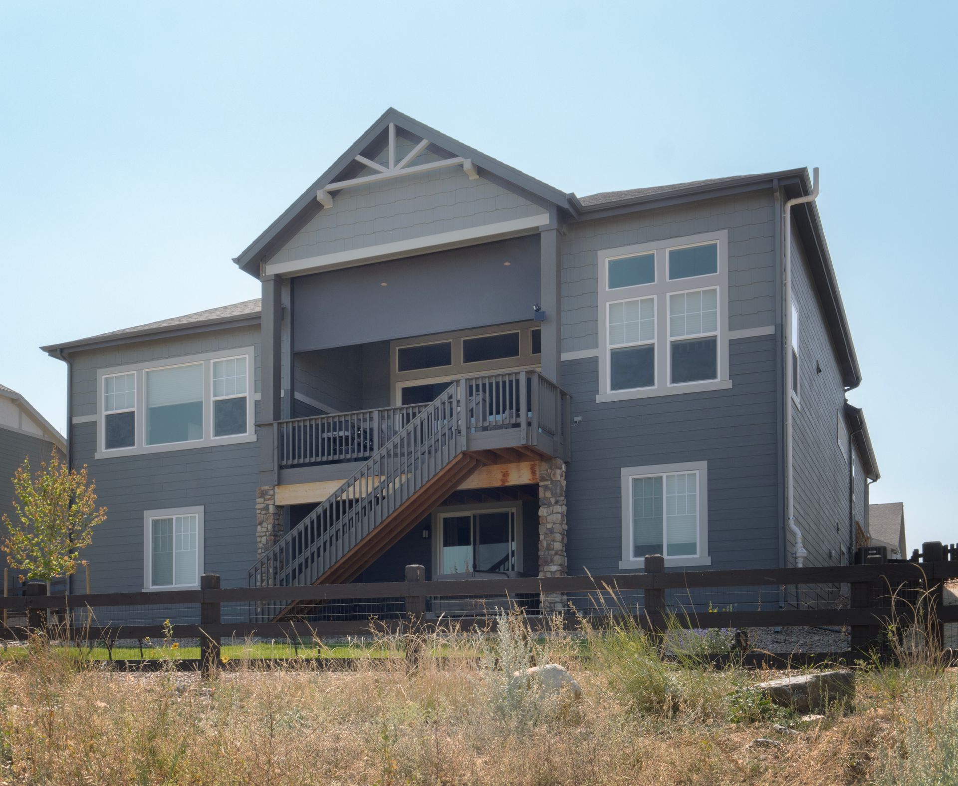 Two-story gray house with a front staircase and balcony behind a fence, in a grassy lot