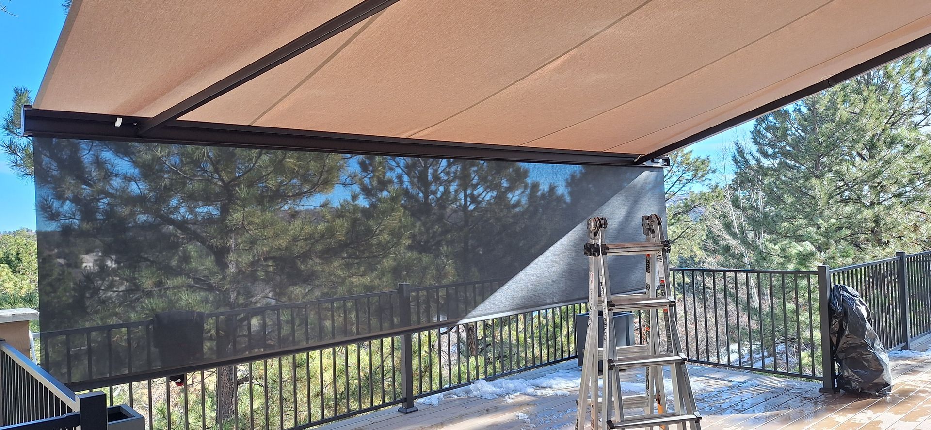 Patio with tan canopy, black railings, folded chair, and scenic trees beyond the deck