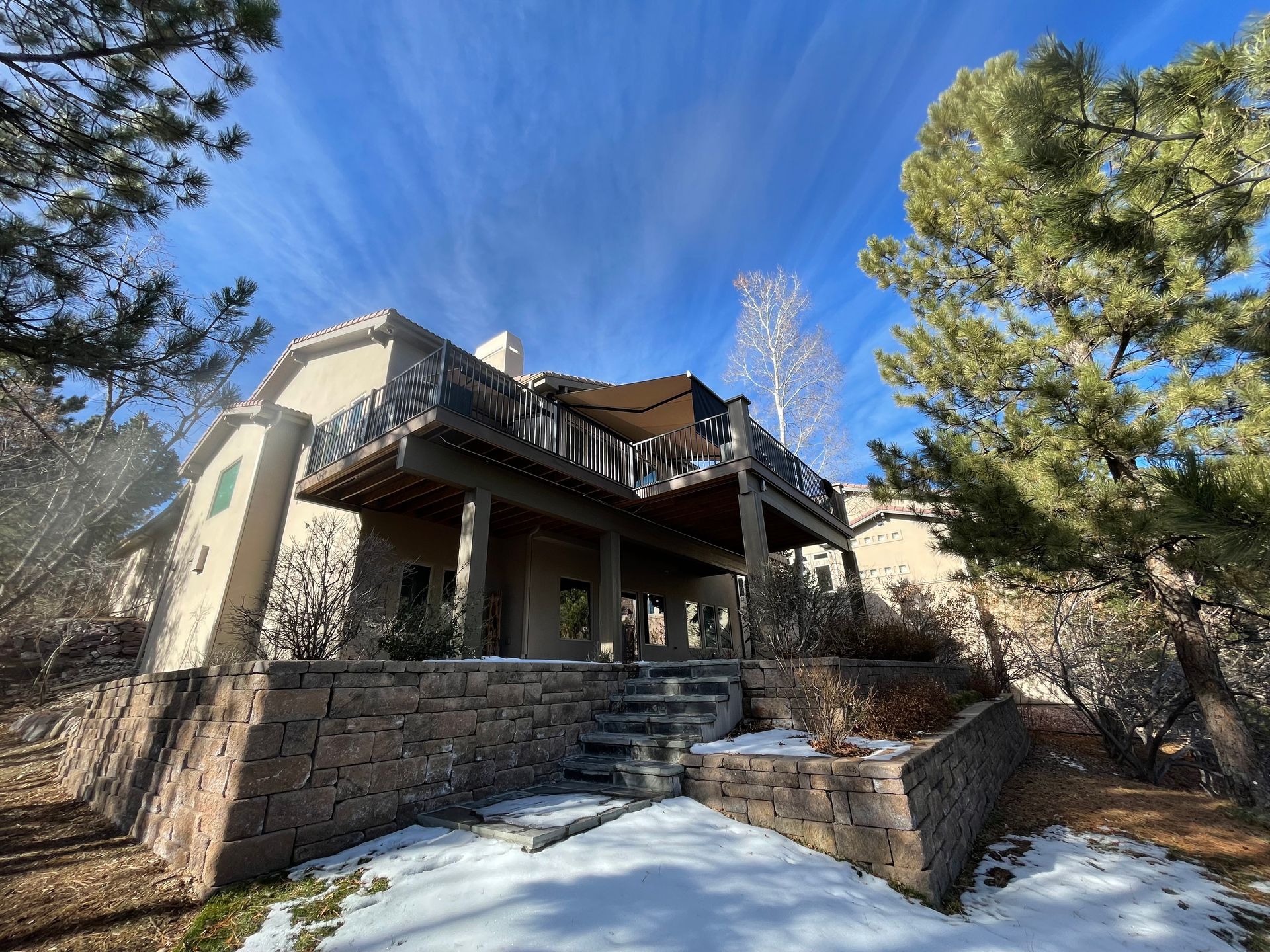 Stone house with a covered porch, front steps, and tall pine trees under a bright blue sky.