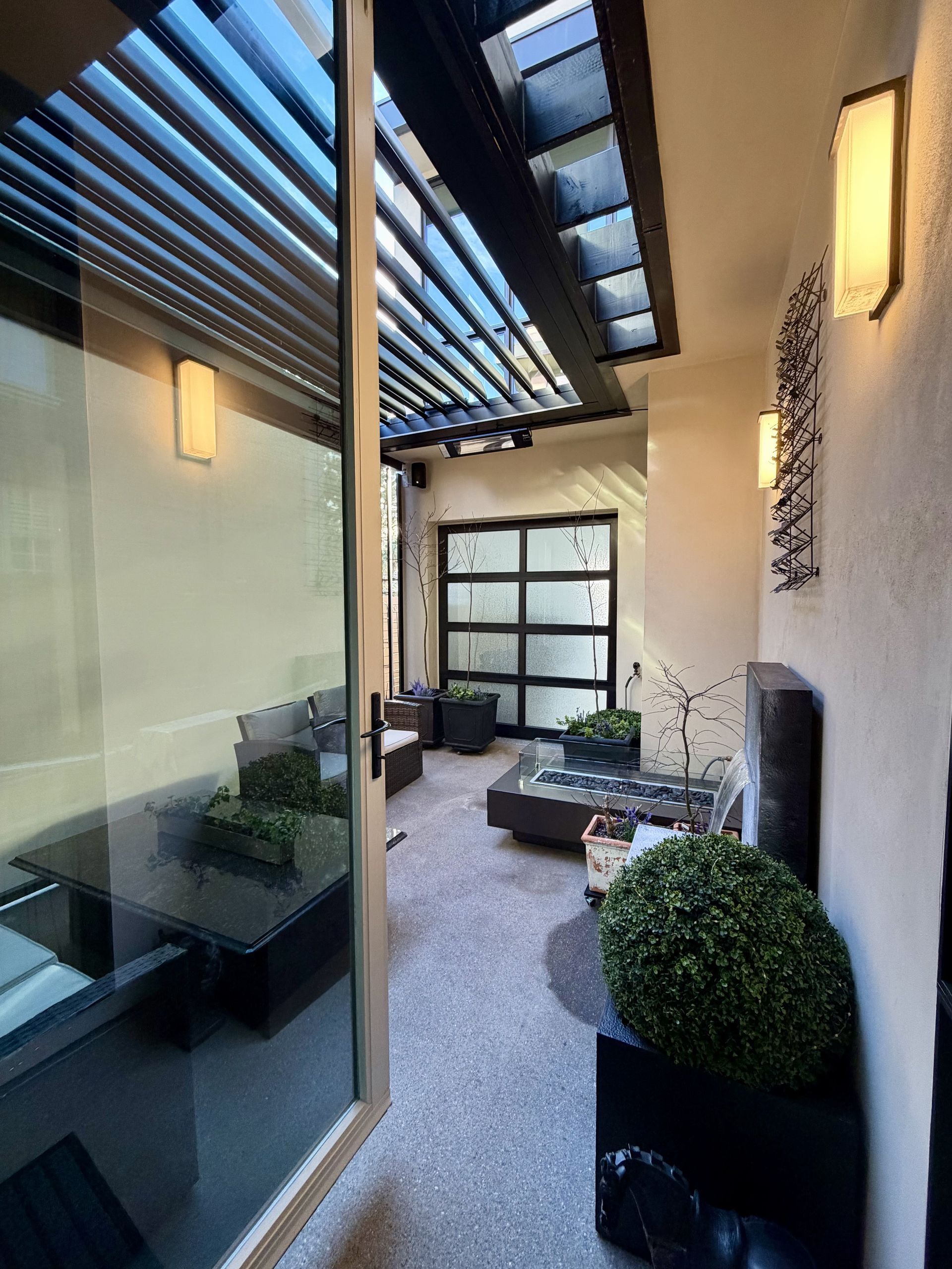 Covered outdoor patio with glass door, black planters, lounge chairs, and wall lights along a narrow walkway