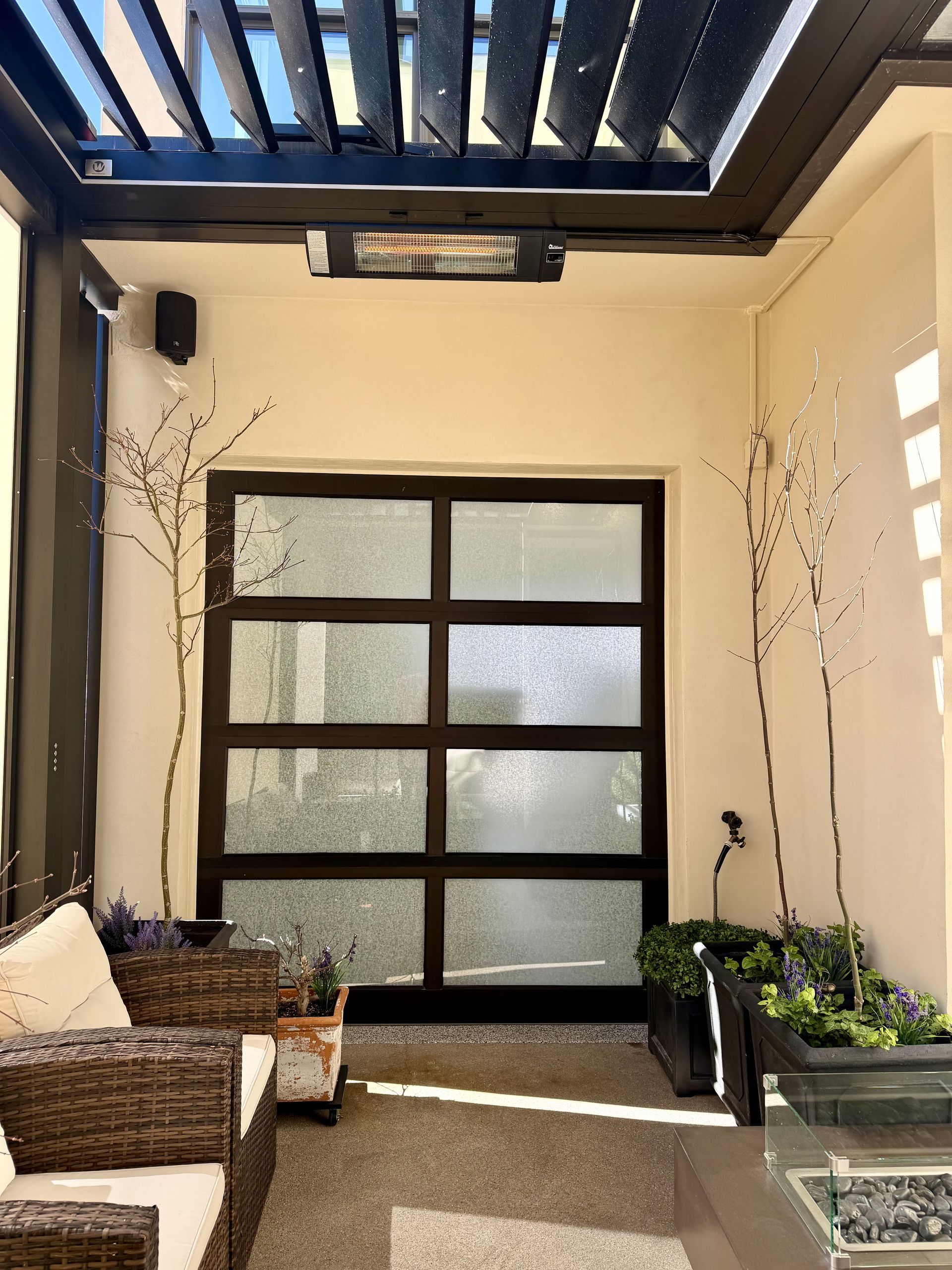 Outdoor patio with wicker chairs, potted plants, and a dark-framed glass door under a pergola