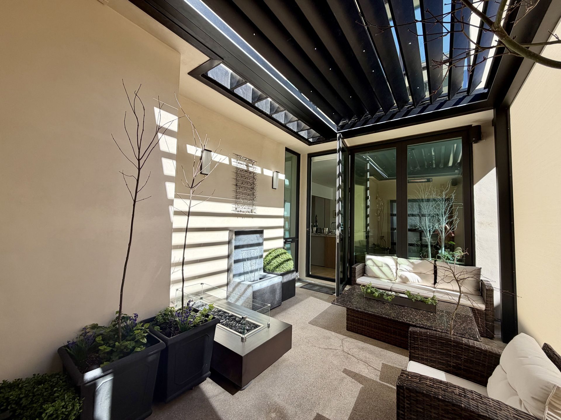 Covered patio with striped canopy, potted plants, and outdoor seating beside a glass wall